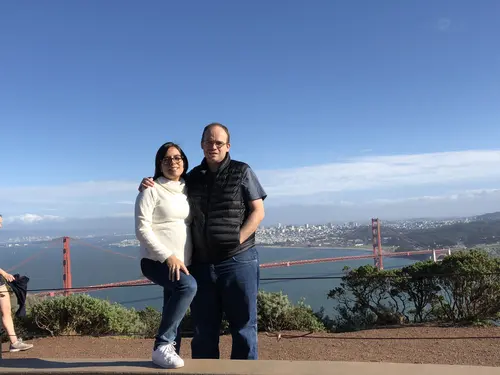 Esteban Rey y su pareja posan en un mirador con un puente rojo al fondo.